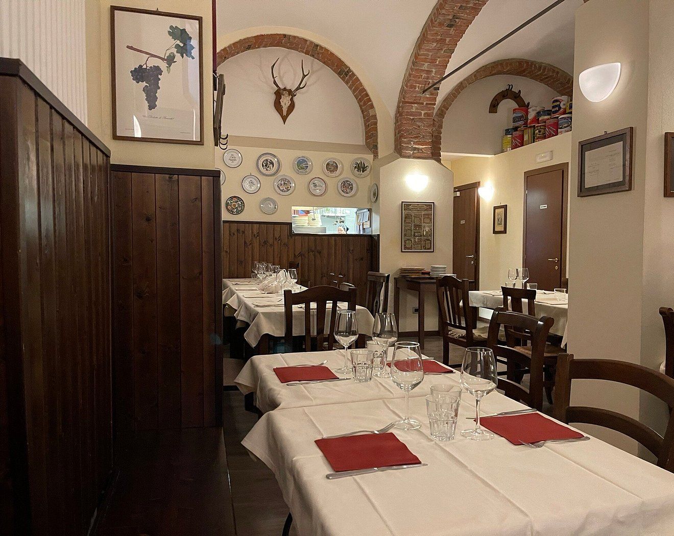 Sala con tovaglie bianche, boiserie in legno scuro, archi in mattoni, un trofeo di corna e barattoli d’epoca in alto