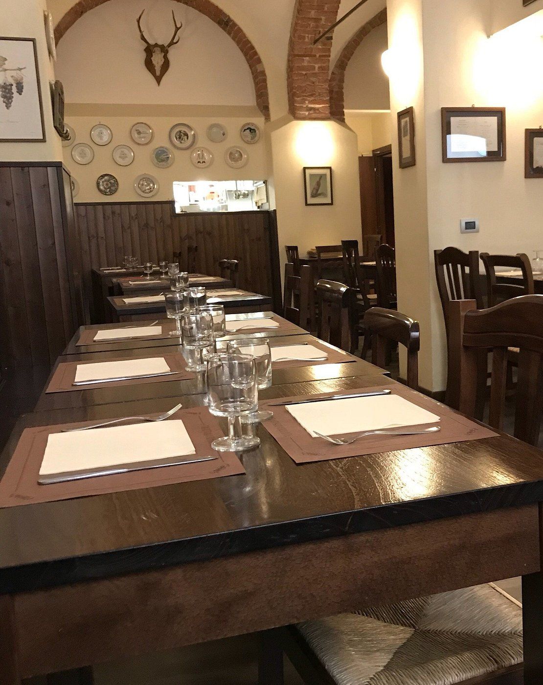 La sala della Gallinaccia: tavoli in legno scuro apparecchiati, archi in mattoni, piatti decorati alle pareti e la finestrella sulla cucina